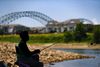 A woman sits in a folding chair fishing. Apartment buildings are seen in the background