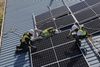 A crew in safety vests and hard hats work on a roof to install solar panels