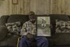 A man in a flannel and jeans sits on a couch holding a framed painting of himself depicted sitting on a levee 