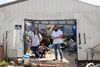 A man and woman in jeans and white t-shirts stand in the doorway of a damaged white garage with piles of stuff on the inside
