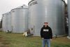 A man in a hoodie and baseball hat stands in front of three silos