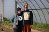 Two people pose with their arms around each other in a hoop house, one with blue and purple hair and a denim jacket covered in buttons, and the other with blue glasses and quilted sweater