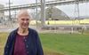 An older woman wearing a gold cross necklace smiles as she stands in front of an industrial plant
