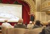 A man speaks into a microphone while he references a slide on a projector screen of historic Memphis along the waterfront. He is presenting to a small audience in an ornate theatre