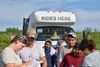 A group of people line up to talk with two women, who are writing on clipboards. In the back is a bus that says "rides here" 