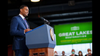 A man in suit and tie stands at a podium, with a green "Great Lakes" sign beside him