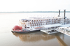 A majestic white riverboat with big red wheels on the front deck and blue details is docked 