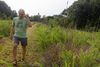 A man wearing a green T-shirt, grey shorts and sandals walks along a path through brush.