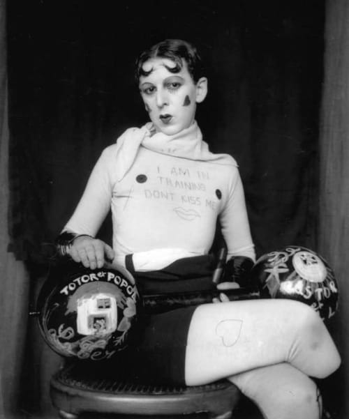 A black-and-white portrait of Cahun dressed as a dandy/mime. His shirt reads "I AM IN TRAINING DON'T KISS ME" with a drawing of lips. Legs crossed, he is poised, madeup, and with a tilt of the head, he gives the camera a knowing, flirty glance. Hearts are drawn on his cheeks and leggings, and he appears alongside what look like bowling balls, painted with words and pictures.