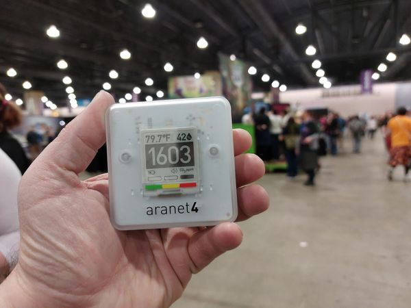 A hand holding an Aranet4 air quality monitor reading 1,603 ppm CO₂ in a convention center expo hall.