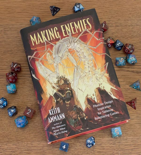 A hardbound copy of Making Enemies on a wooden dining table, surrounded by blue and red polyhedral dice.