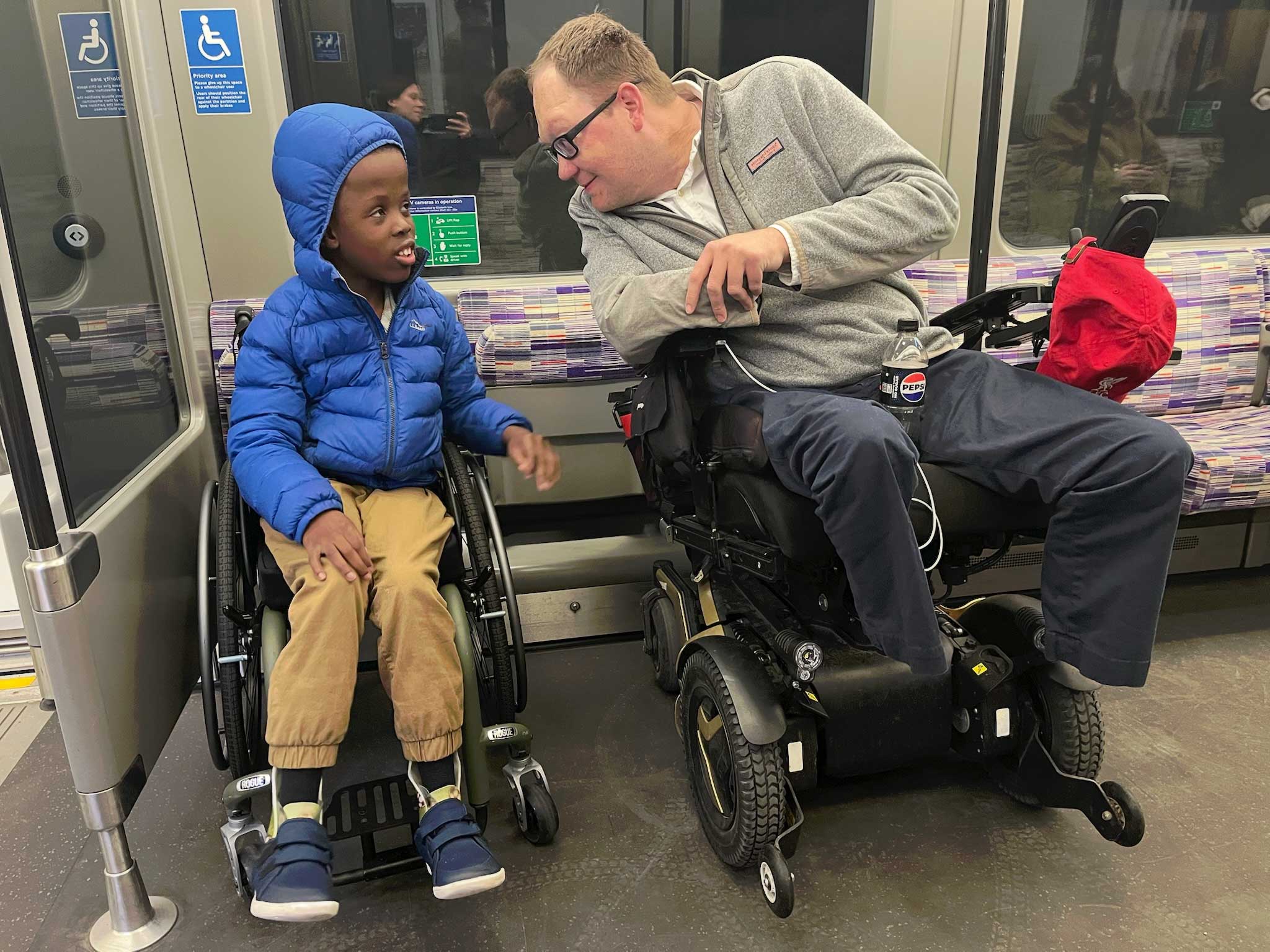 John and Robert seated in their wheelchairs, talking while on a train in London.
