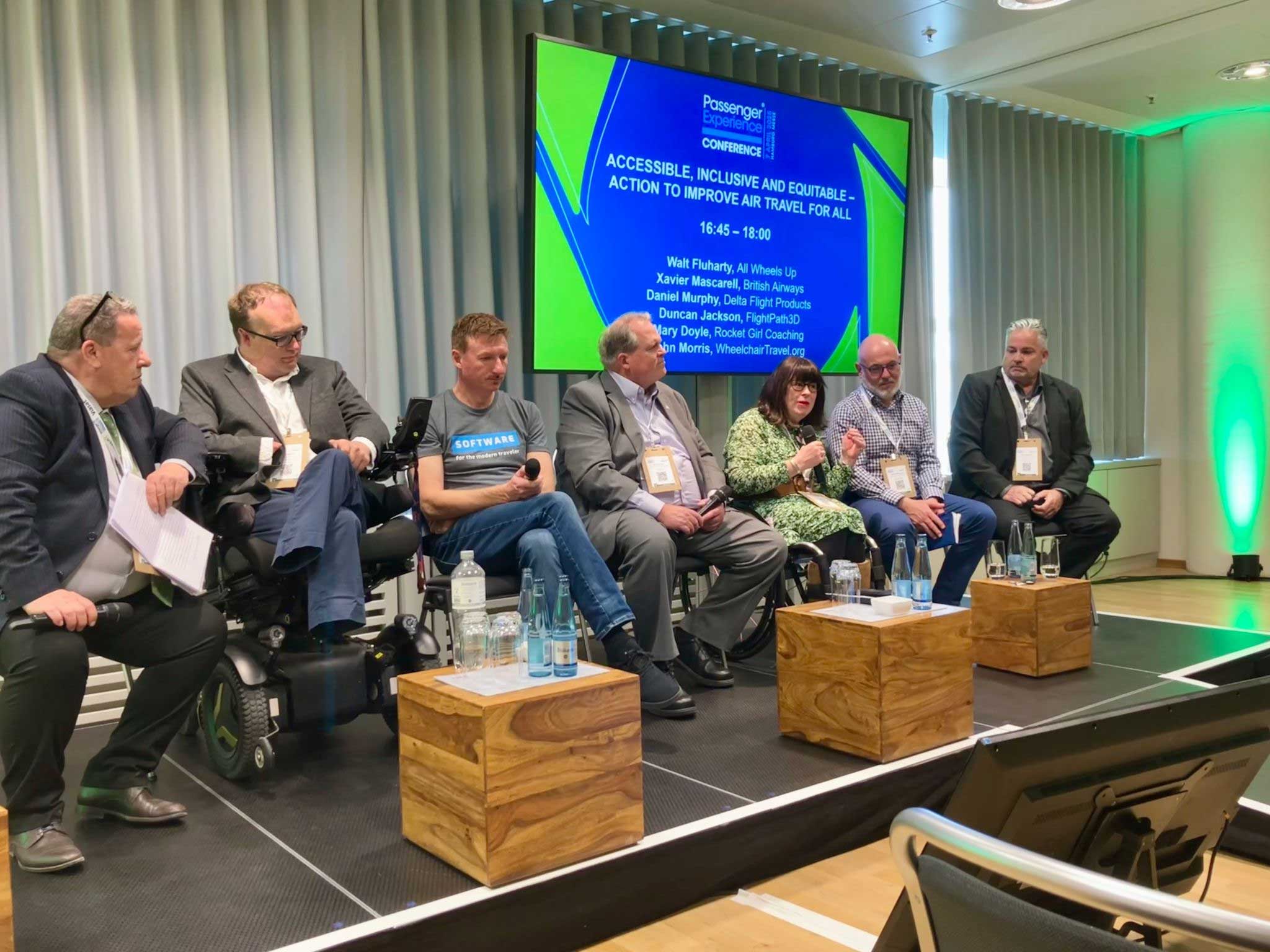 Group of seen speakers on stage, including John and fellow advocate Mary, both seated in their wheelchairs.