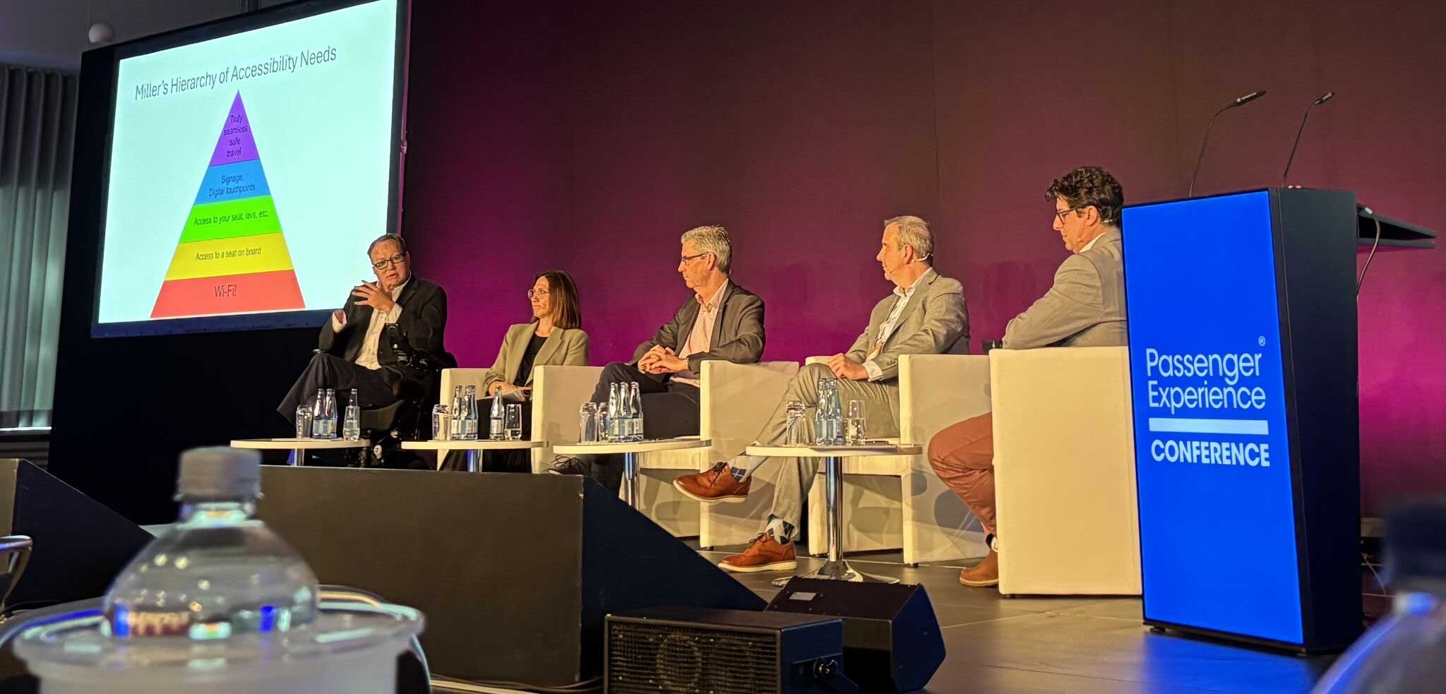 John speaking and using his hands to motion while seated in his wheelchair on stage next to fellow panelists.