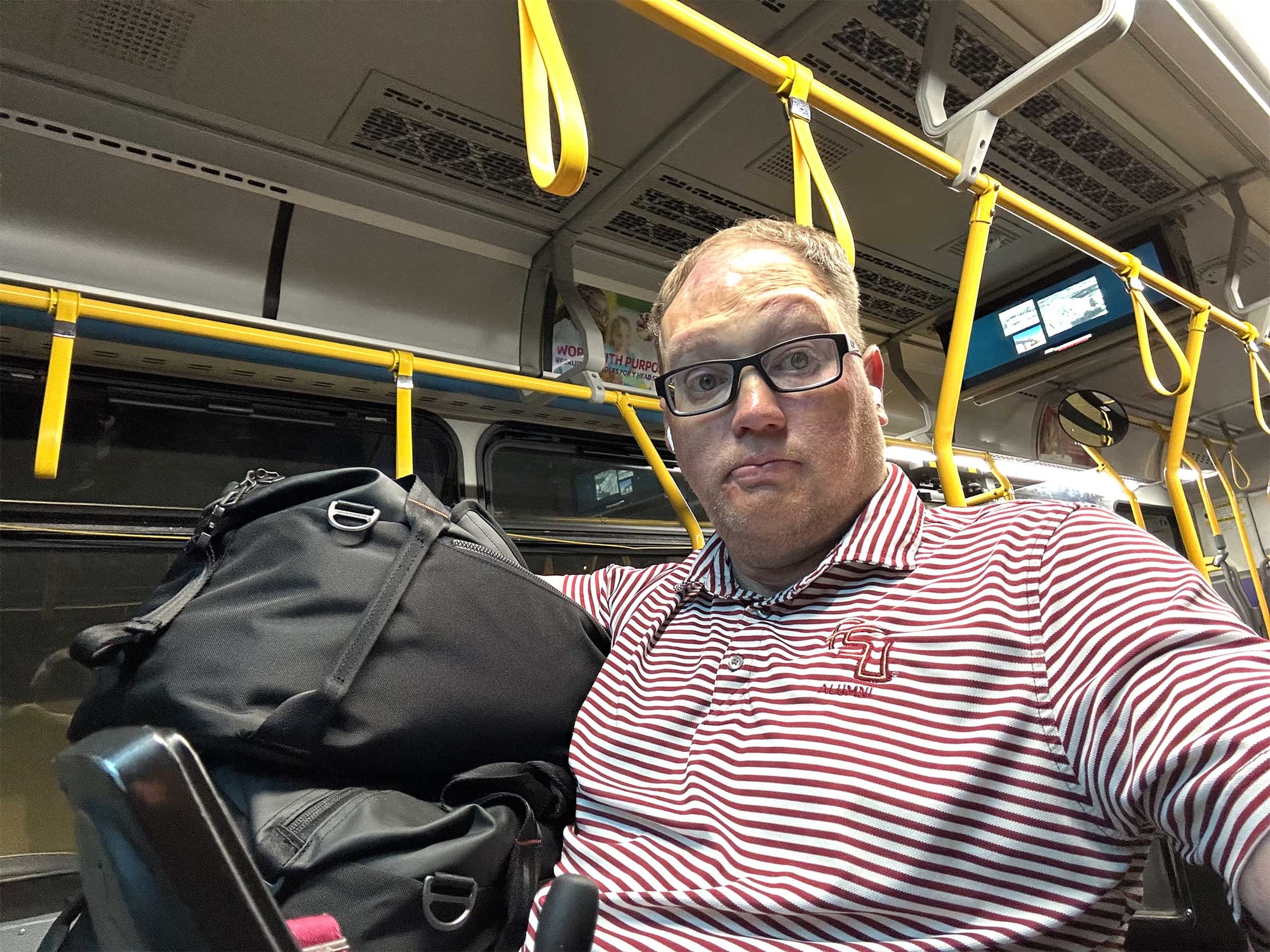 Selfie of John seated in his wheelchair while holding two duffle bags on a city bus.