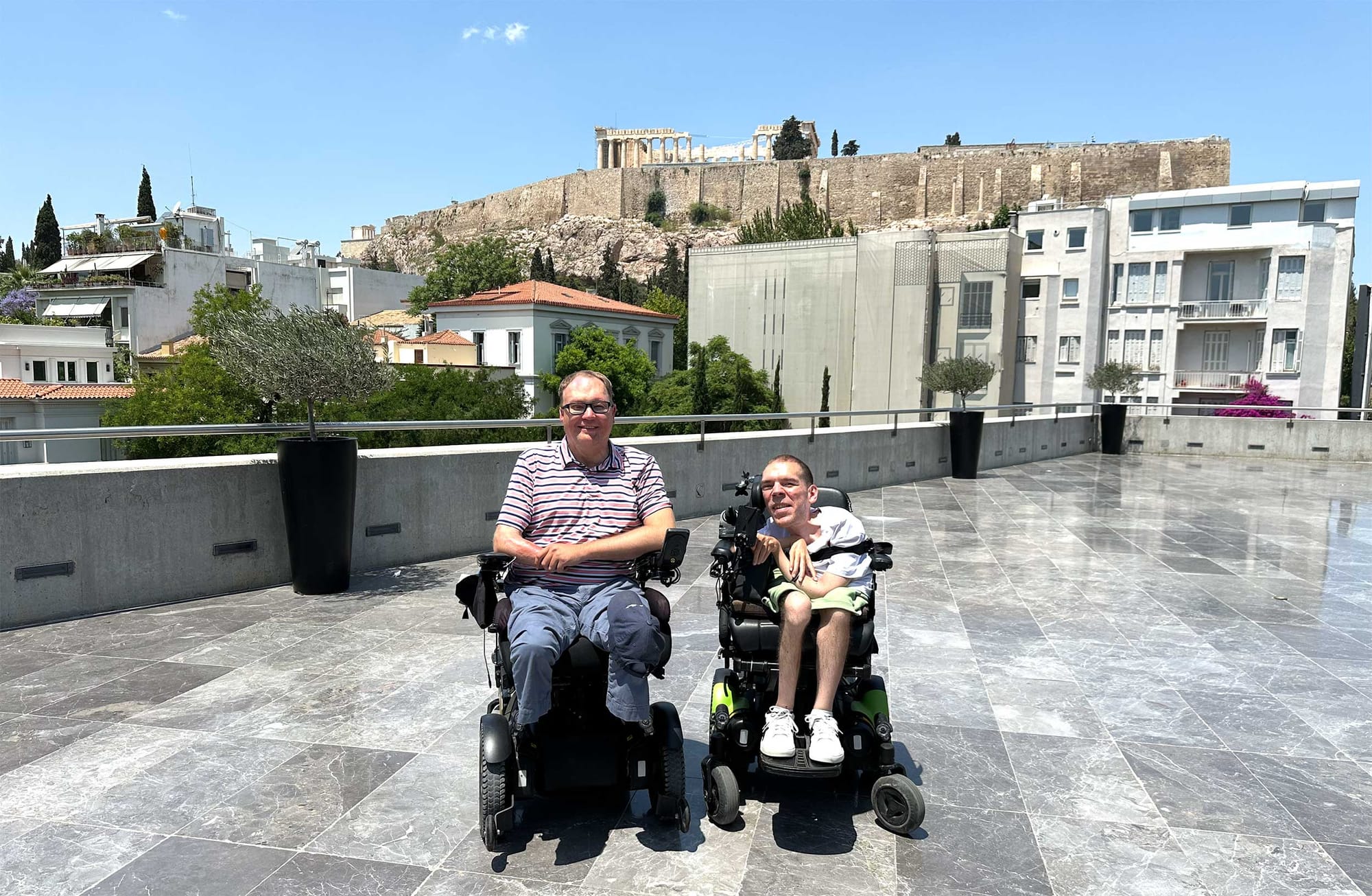 John seated in his wheelchair next to another wheelchair user on the rooftop of a building with the Acropolis seen in the distance.