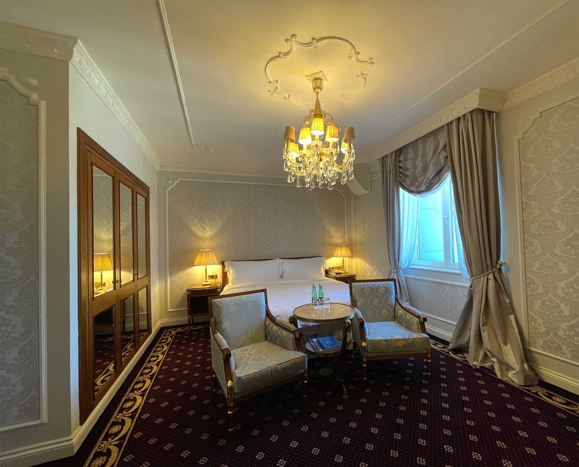 King size bed in room with large chandelier, fine carpet and chairs.