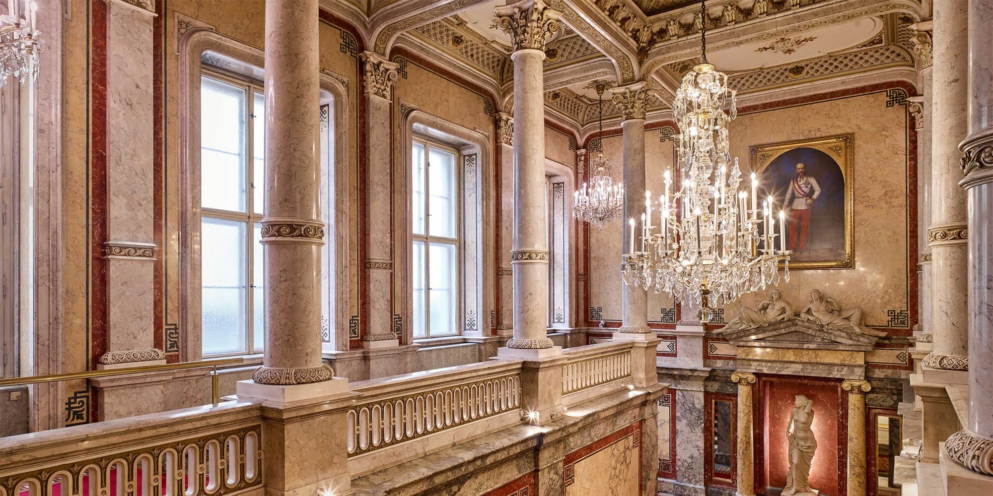 Ornate chandelier over staircase at luxury hotel with marble columns and large windows, and a portrait of a king.