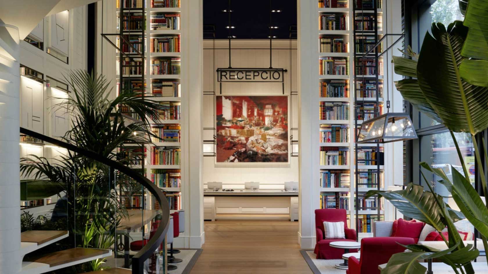 Reception desk at hotel, sitting room with plush seats, plants and floor to ceiling bookshelves.