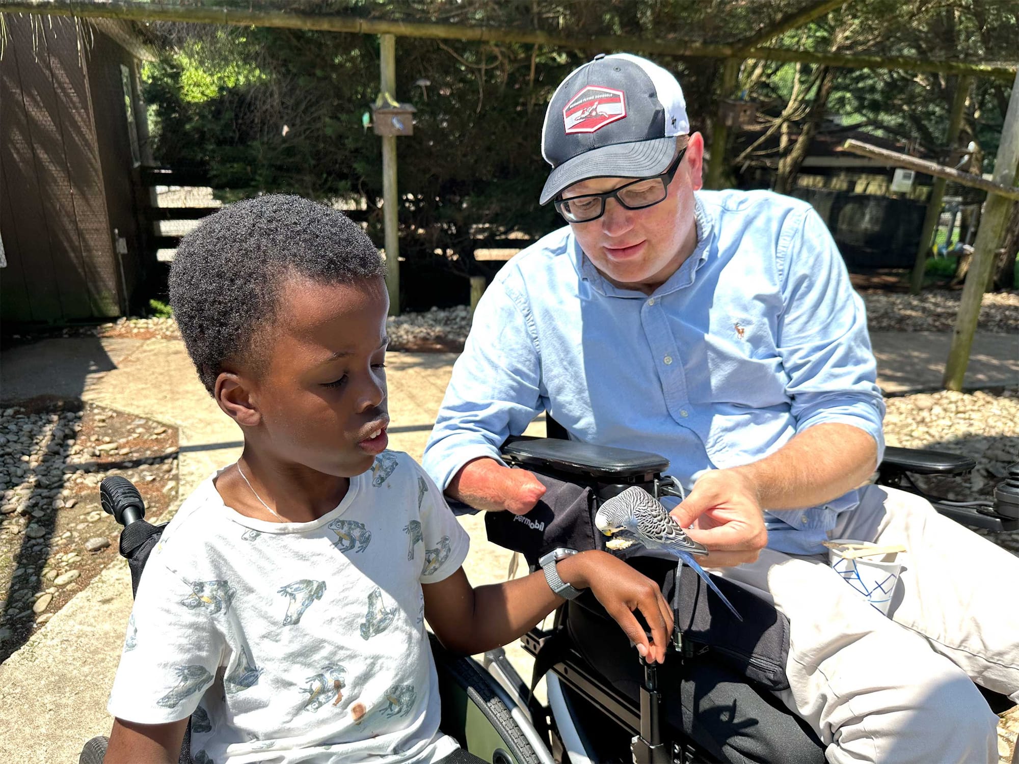 John and Robert seated in their wheelchairs feeding a grey, white and blue bird.