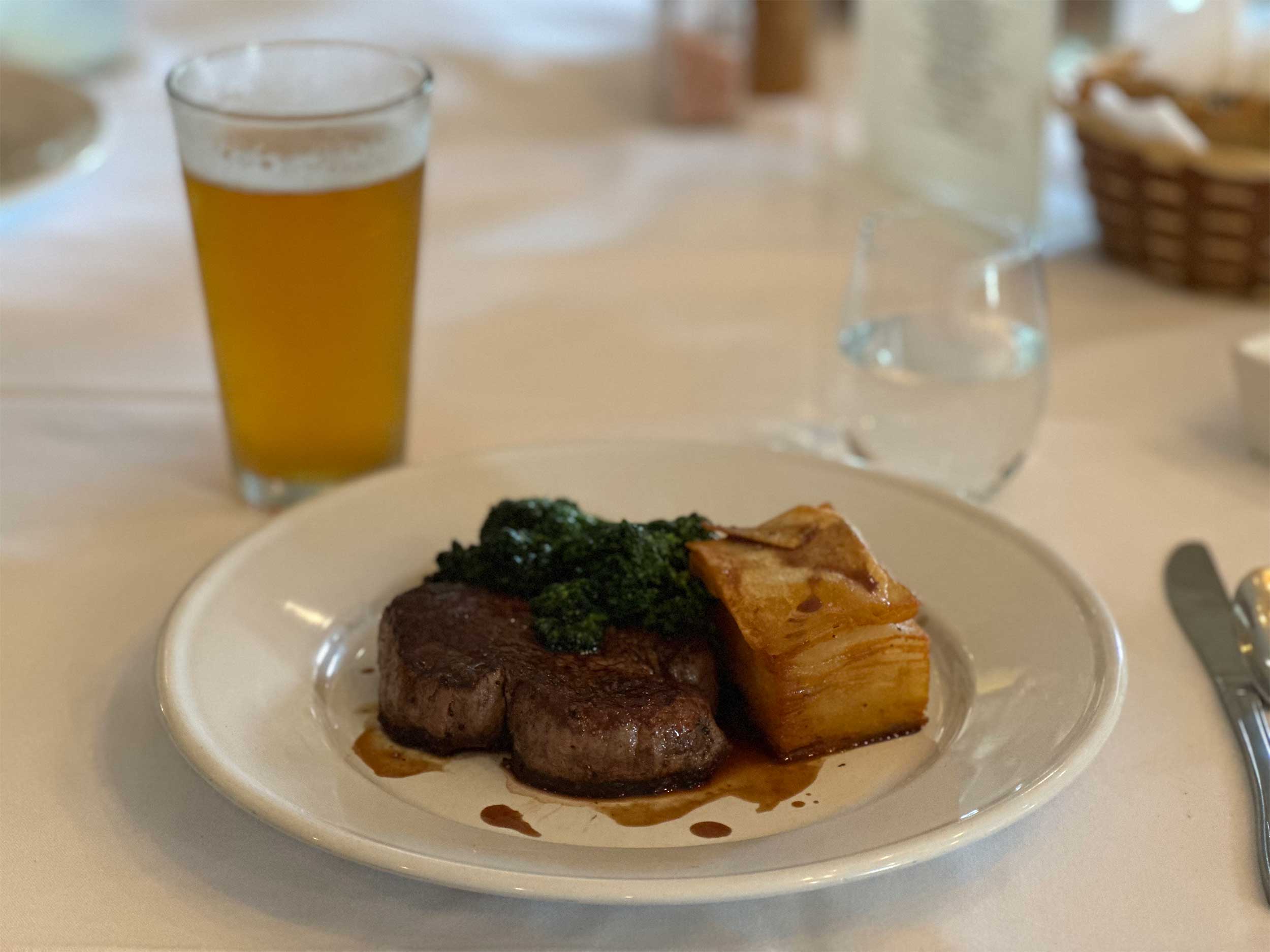 Steak, broccoli and potatoes on a plate, with a beer and bread basket in the background.
