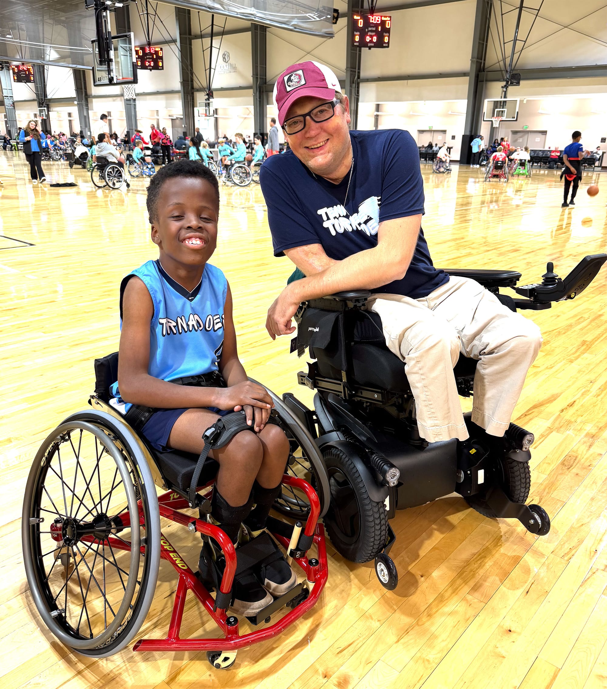 Robert seated in his basketball wheelchair next to John in his power wheelchair on a basketball court.