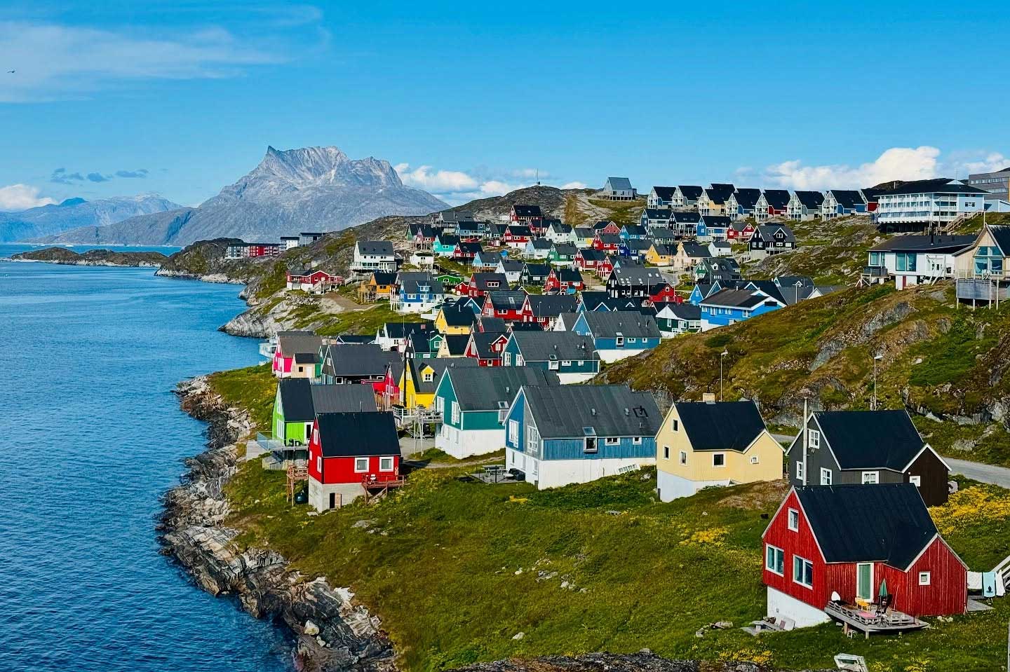 Colorful buildings along Greenland coast, rocky and with grass. Mountain seen in the background.