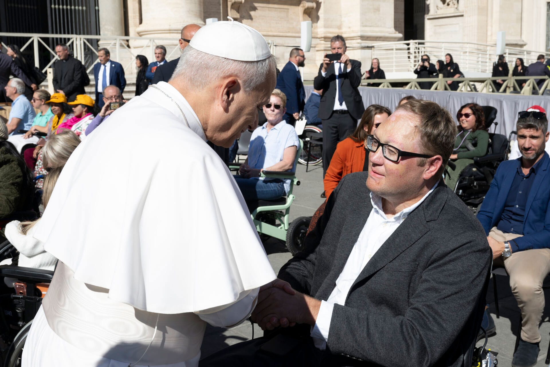 John seated in his wheelchair shaking hands with Pope Leo.