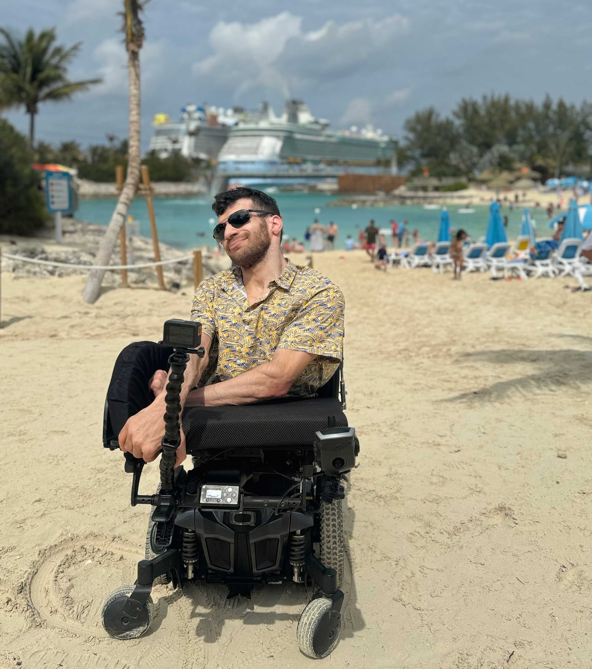 Nolan seated in his wheelchair on a sandy beach with a cruise ship in the background.