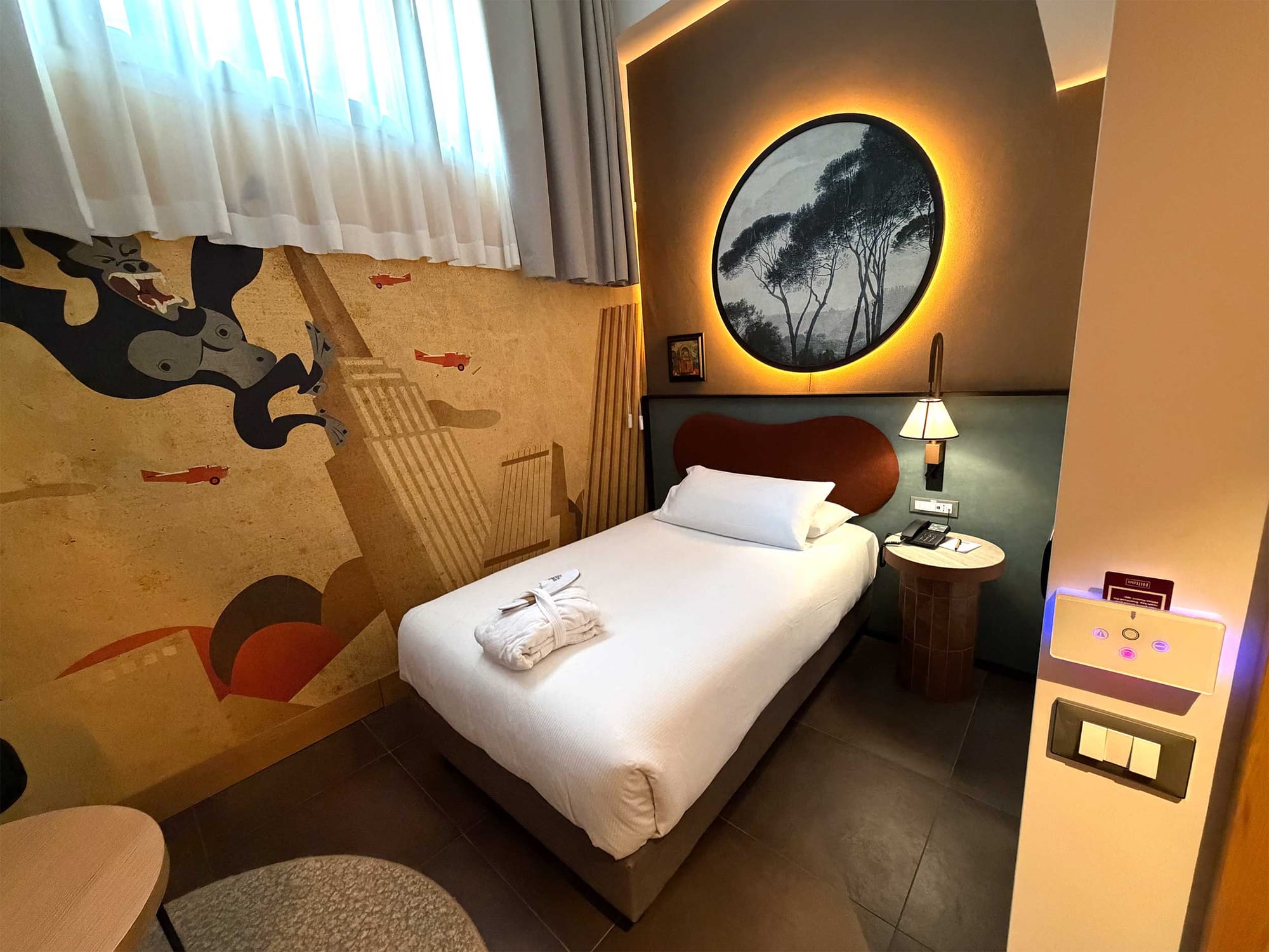 Single bed in compact guest room featuring bold artistic wallpaper, a backlit circular artwork above a leather headboard, and a small nightstand.