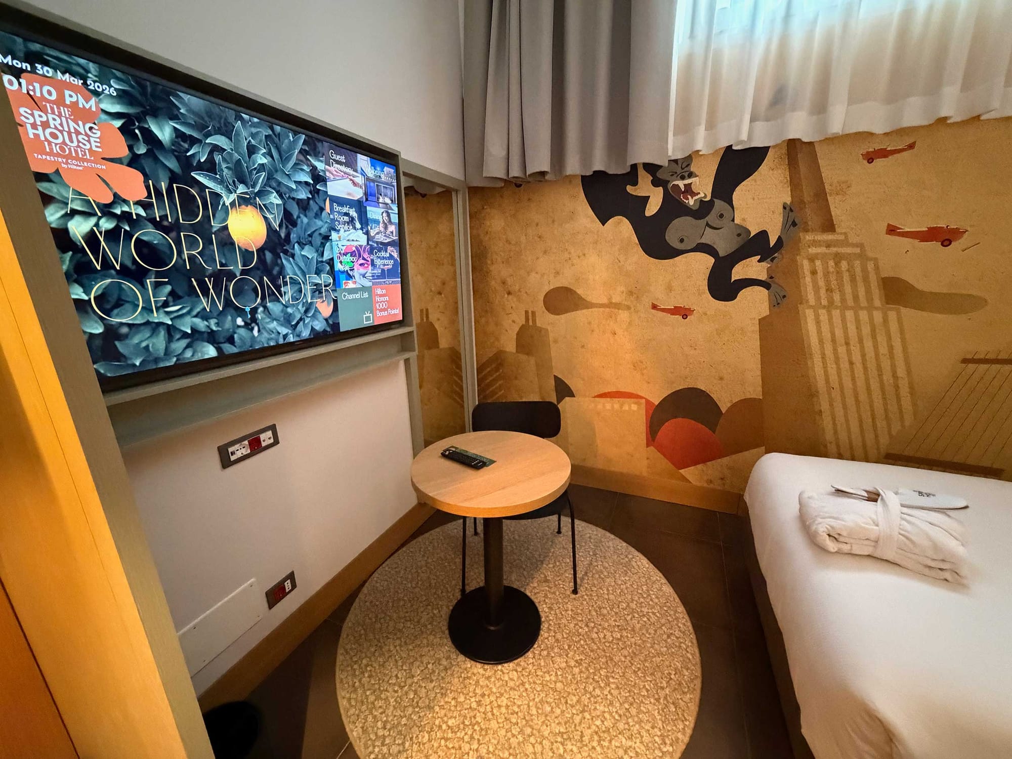 Wall-mounted television, round pedestal table with chair, and artistic mural in compact room showing the limited floor space.