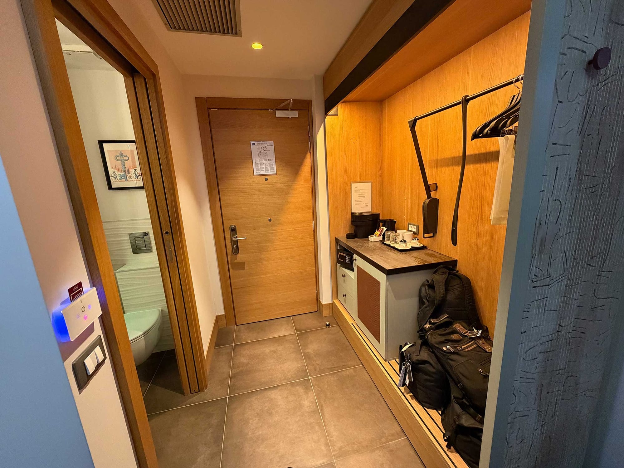 Narrow entry hallway showing the room door, bathroom entrance on the left with the toilet visible, and an open closet area with coffee maker and hanging rod on the right.