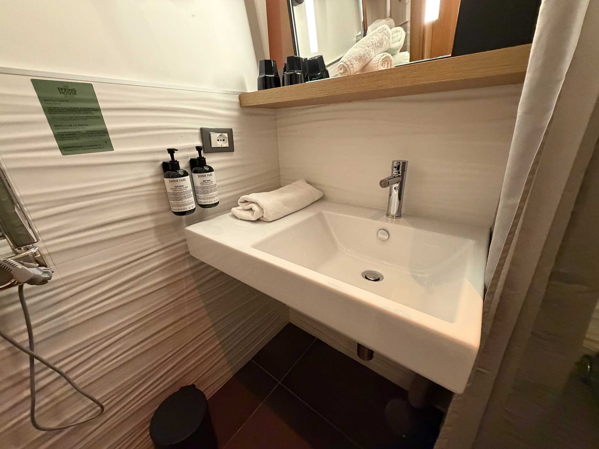 Close-up of wall-mounted sink with single faucet, shelf with mirror above, soap dispensers, and exposed plumbing underneath.