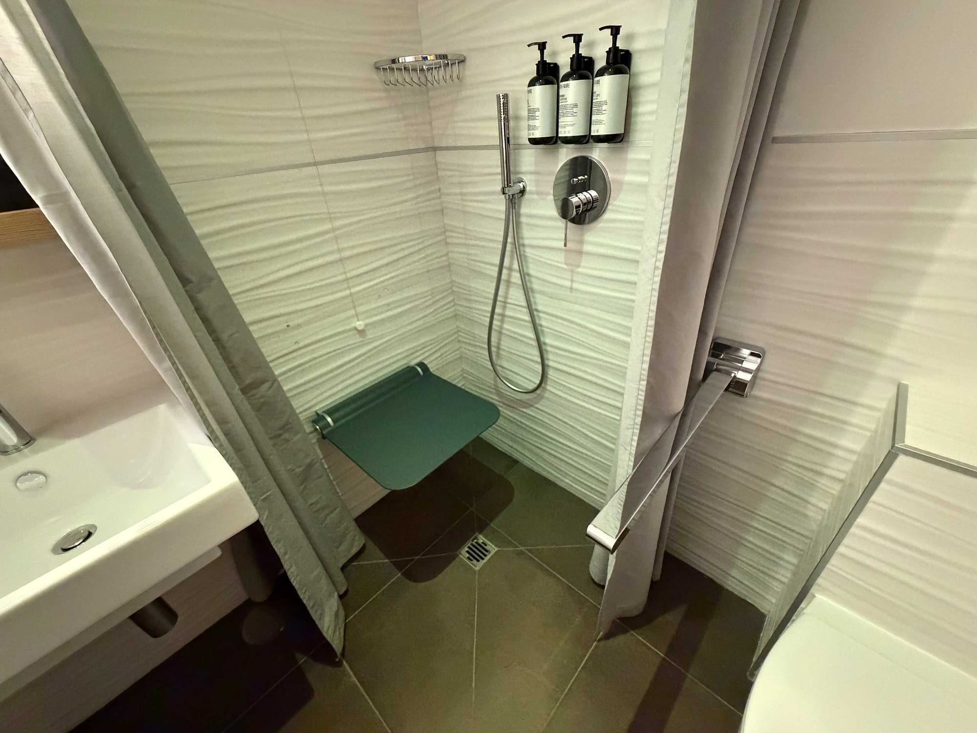 Roll-in shower with a teal fold-down seat, rain shower head, handheld shower, wall-mounted soap dispensers, and shower curtain — with no grab bars visible on any wall.