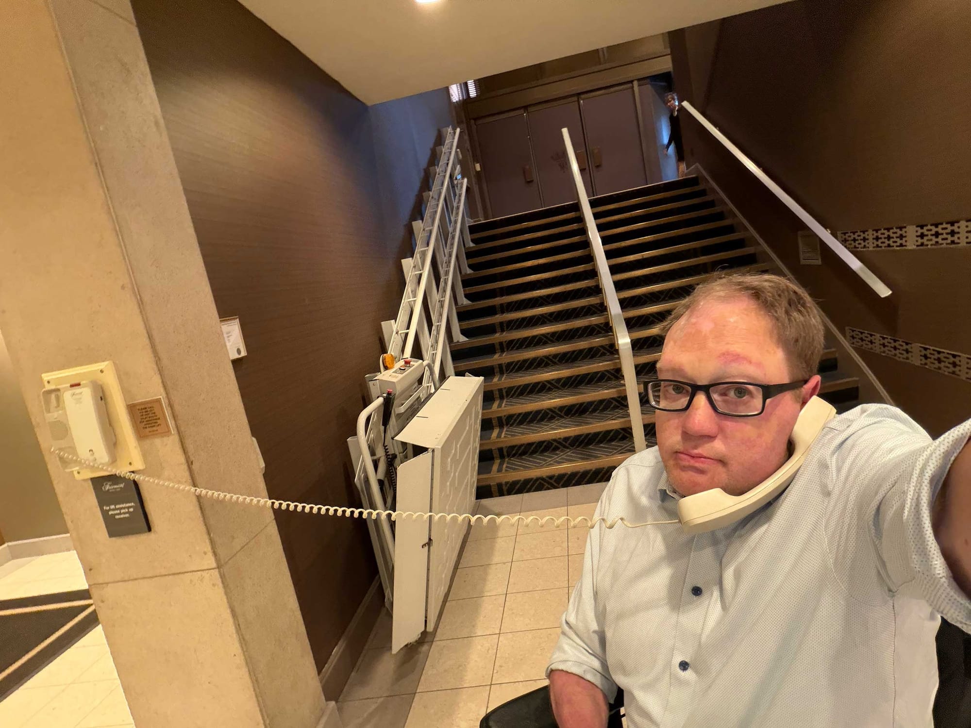 John taking a selfie while holding a phone to his ear in front of a wheelchair stair lift.