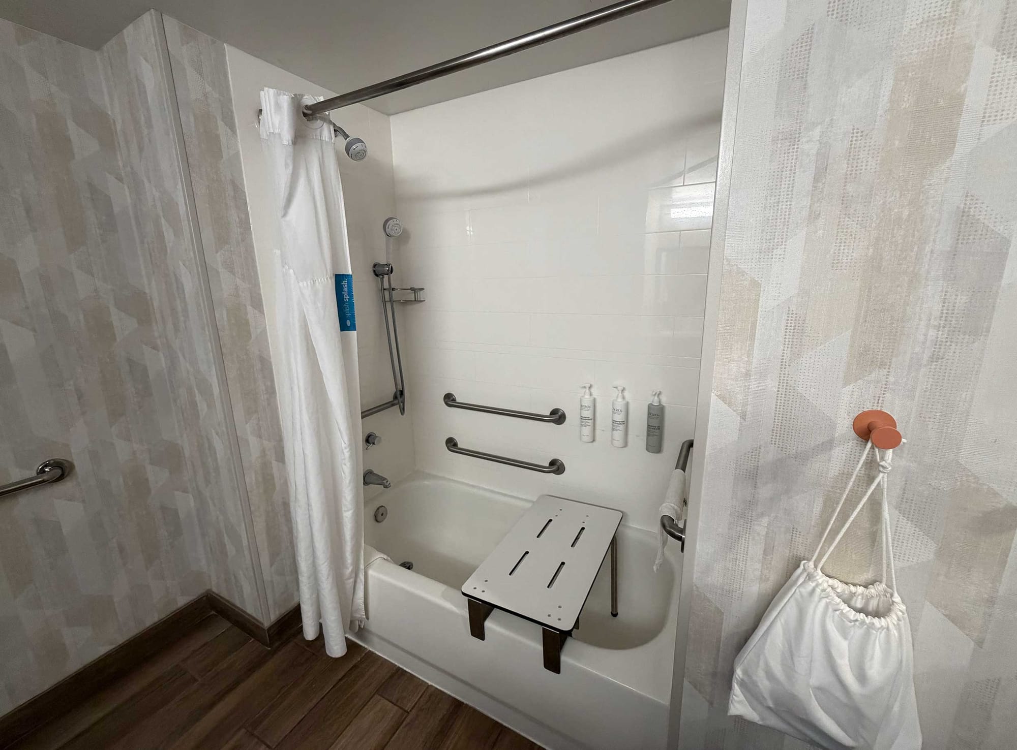 Accessible bathtub with a removable ADA-compliant bathtub seat installed spanning the tub width, multiple grab bars, a handheld shower head and wall-mounted soap dispensers.