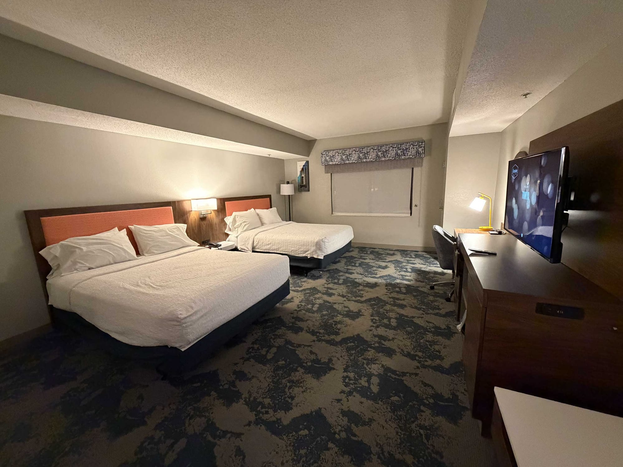 Two queen beds in a spacious room with carpet, a television on the dresser and a desk area in the background.