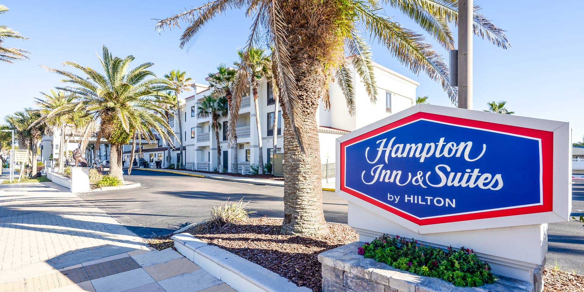 Hampton Inn and Suites sign in front of hotel with palm trees and sidewalk.