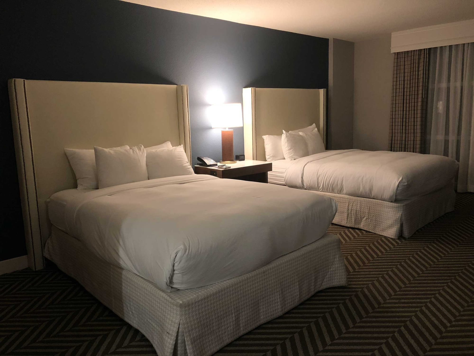 Two queen beds with tall cream upholstered headboards against a dark accent wall, with herringbone patterned carpet.