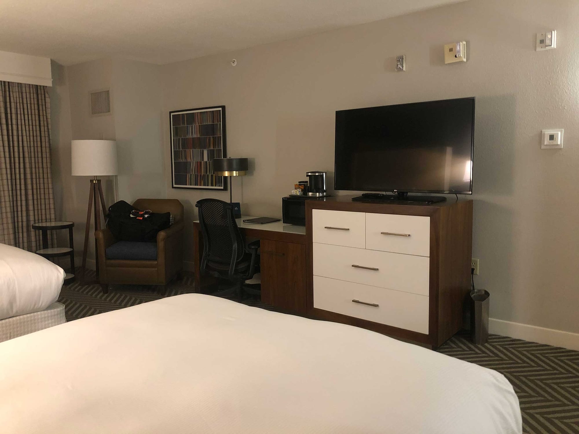 Desk area with ergonomic office chair, armchair, dresser with white drawer fronts, wall-mounted television and coffee maker.
