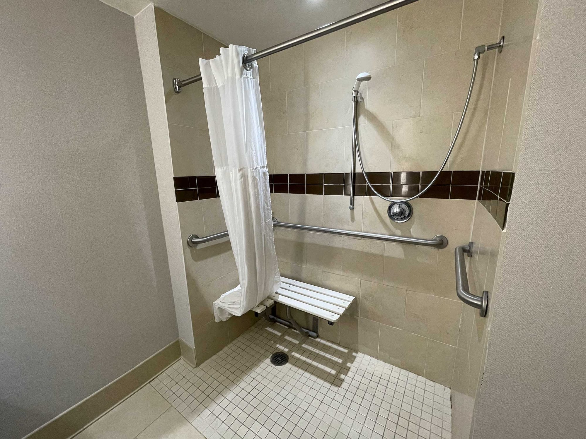 Roll-in shower with a fold-down seat affixed to the back wall rather than a side wall, with grab bars on the side walls and a handheld shower head.