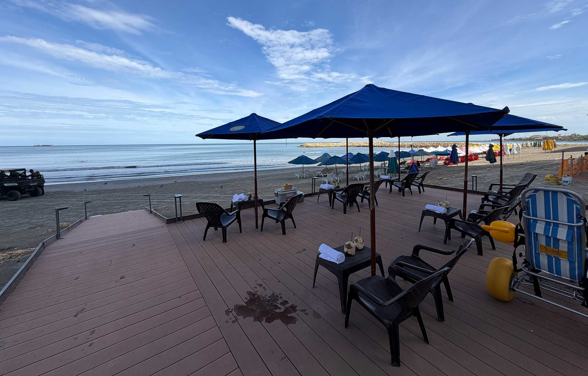 Wooden boardwalk deck with blue umbrellas, black chairs and side tables holding rolled towels and fresh coconuts, overlooking the sea at Cartagena Beach.