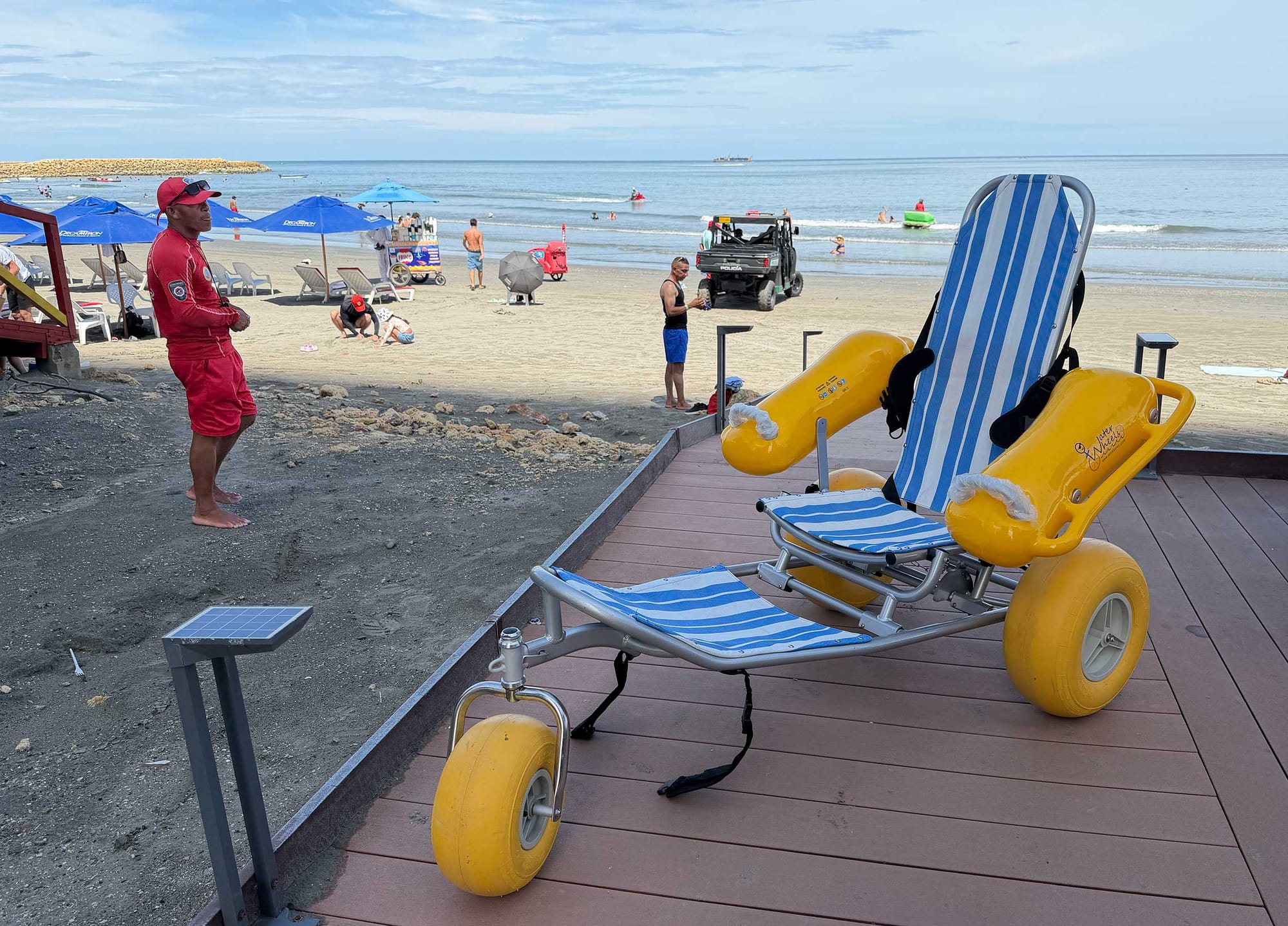Blue and white striped WaterWheels floating beach wheelchair with large yellow balloon tires and flotation armrests, parked on the boardwalk deck as a lifeguard in red stands on the sand nearby.