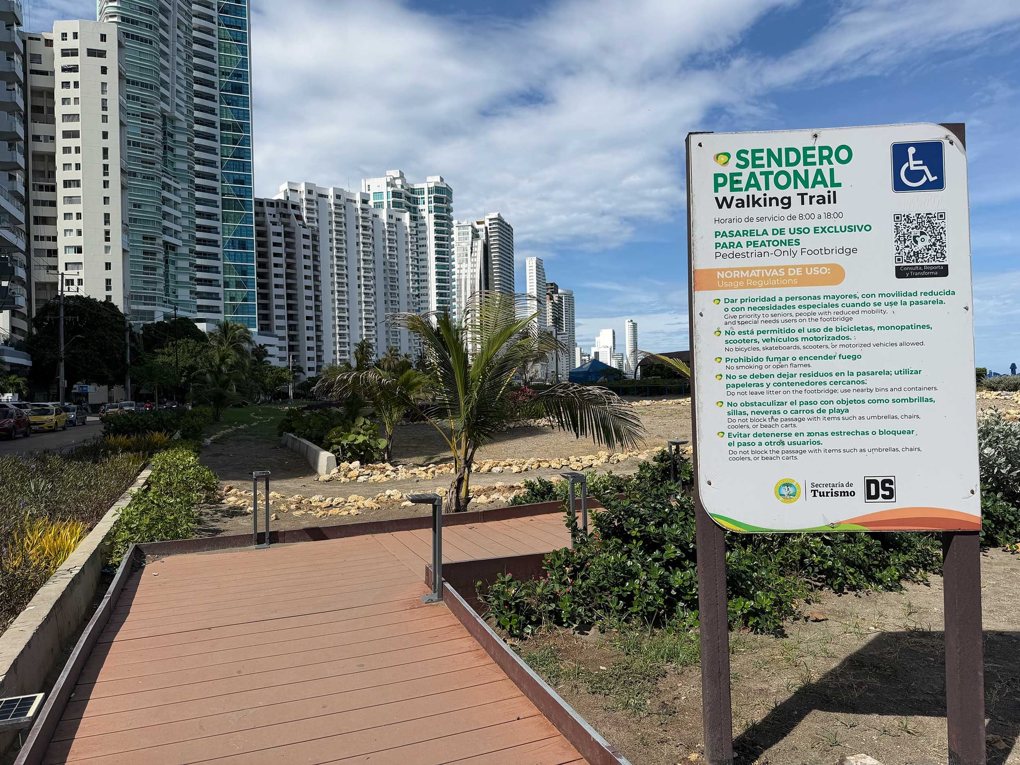 Entrance ramp to the walking trail with a posted sign displaying the wheelchair accessibility symbol and usage regulations in Spanish and English, backed by tall buildings.