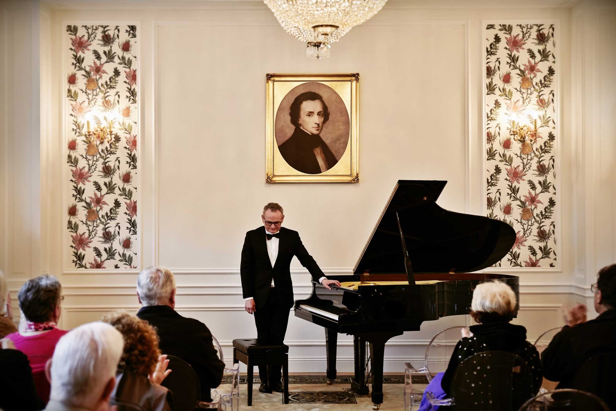 Pianist in front of a piano with a picture of Chopin hanging on the wall behind him.