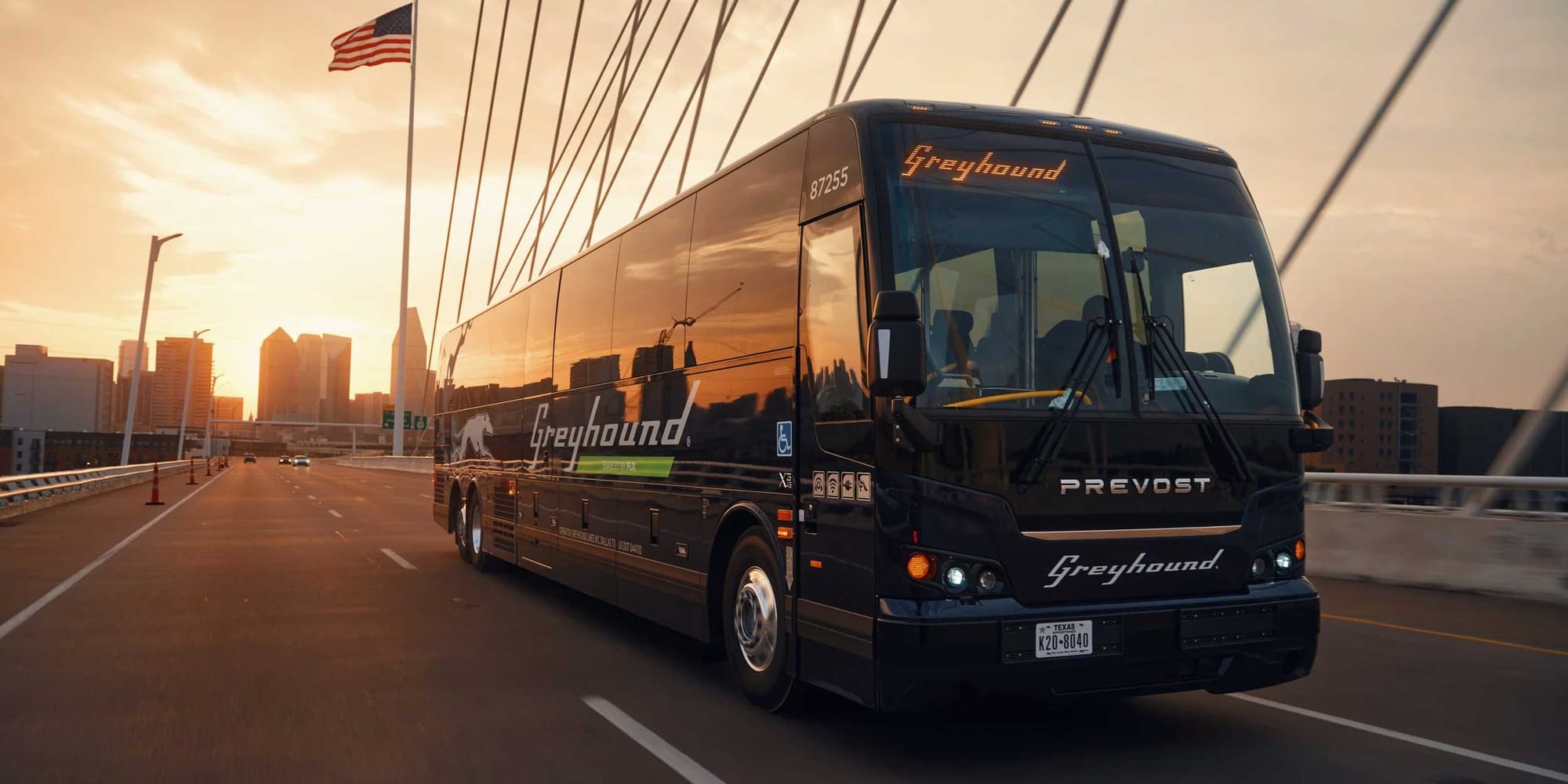 Greyhound bus on highway bridge with American flag and city skyline in the background.