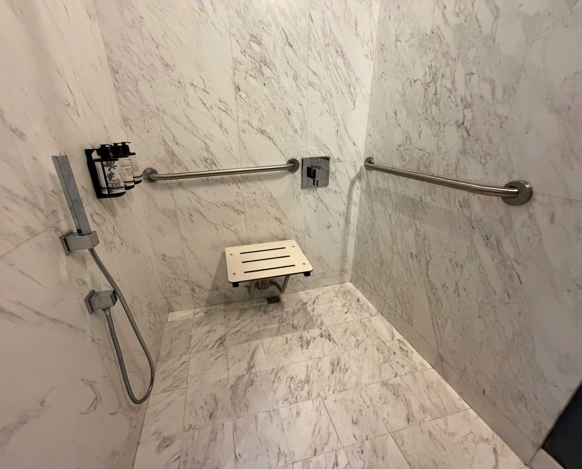 Roll-in shower with a small fold-down seat mounted at the center of the back wall, a single horizontal grab bar on the right side wall positioned far from the seat, a handheld shower head mounted on the left wall, and two wall-mounted dispensers.