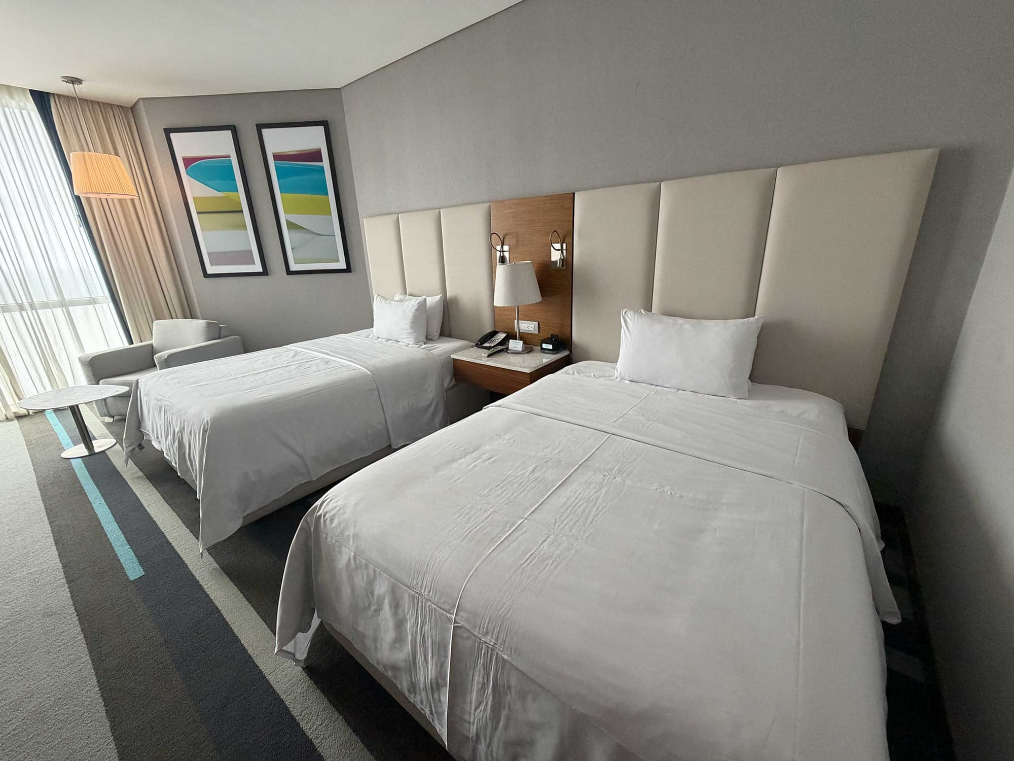 Guest room with two queen beds on solid platform bases, a wall-mounted television, desk with office chair and floor-to-ceiling windows with sheer curtains.