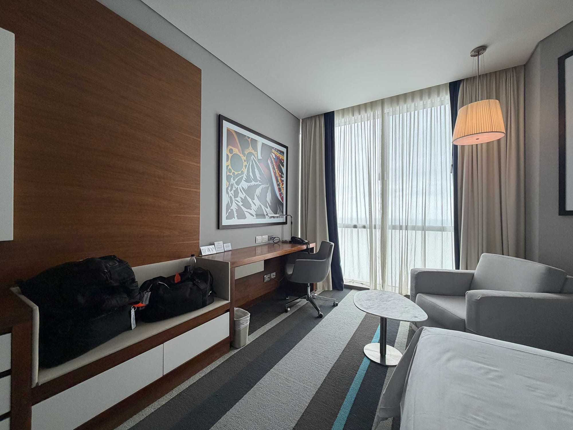 Wall opposite beds with wood-paneled desk and dresser unit with a roll-under opening, an office chair, a sofa, a small round side table.