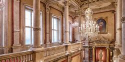 Ornate chandelier over staircase at luxury hotel with marble columns and large windows, and a portrait of a king.